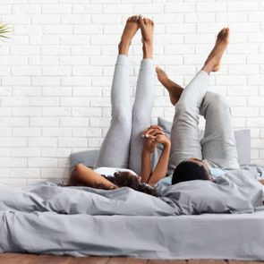 Couple in love. Young happy african-american man and woman lying on bed at home raising legs up, lazy family resting on weekend, copy space