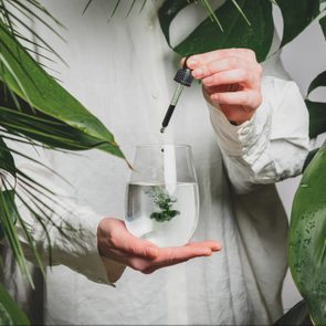 woman in a white shirt adds drops of chlorophyll to glass of water standing next to palm trees