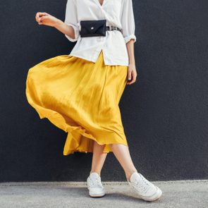 Horizontal cropped image of stylish slim woman in beautiful yellow skirt. Caucasian female fashion model standing over gray wall background outdoor with copy space.